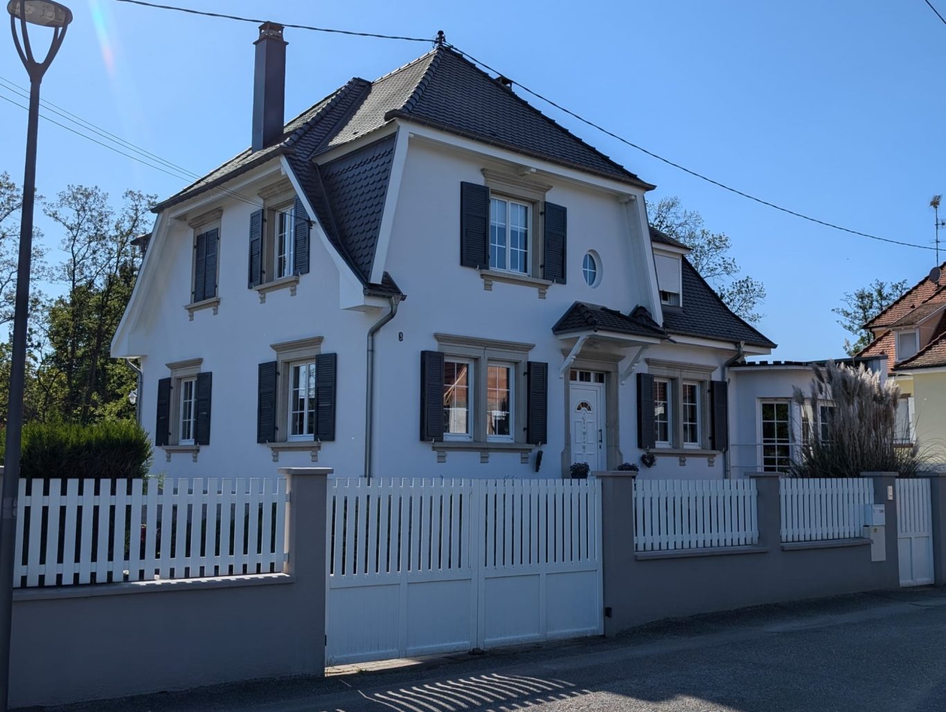Après - Facade grande maison Maison blanche avec volets, clôturée, située sous un ciel bleu ensoleillé.